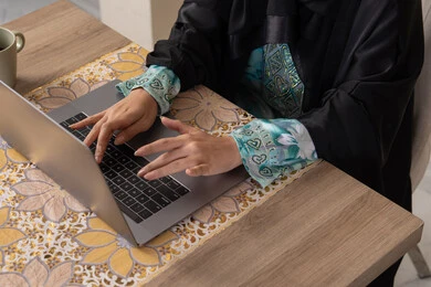 Enjoying coffee, integrating modern technology with daily routine, healthy lifestyle, Saudi Arabian Gulf woman wearing abaya and hijab sitting at a table at home using laptop, Emirati woman putting on headphones, calm morning atmosphere at home