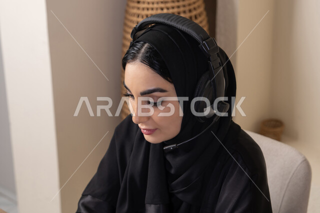 أجواء صباحية هادئة في المنزل، الاستمتاع بشرب القهوة، دمج التكنولوجيا الحديثة مع الروتين اليومي، نمط حياة صحي، امراه عربية خليجية سعودية ترتدي العباءة والحجاب تجلس على طاولة في المنزل تستخدم الحاسب المحمول، سيدة اماراتية تضع الهيدفون