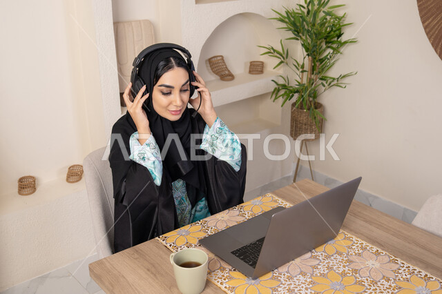 الاستمتاع بشرب القهوة، دمج التكنولوجيا الحديثة مع الروتين اليومي، نمط حياة صحي، امراه عربية خليجية سعودية ترتدي العباءة والحجاب تجلس على طاولة في المنزل تستخدم الحاسب المحمول، سيدة اماراتية تضع الهيدفون ، أجواء صباحية هادئة في المنزل