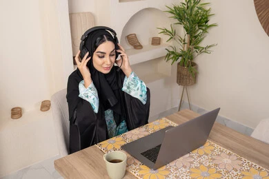 Enjoying coffee, integrating modern technology with daily routine, healthy lifestyle, Saudi Arabian Gulf woman wearing abaya and hijab sitting at a table at home using laptop, Emirati woman putting on headphones, calm morning atmosphere at home