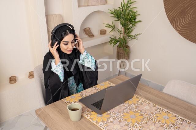 أجواء صباحية هادئة في المنزل، الاستمتاع بشرب القهوة، دمج التكنولوجيا الحديثة مع الروتين اليومي، نمط حياة صحي، امراه عربية خليجية سعودية ترتدي العباءة والحجاب تجلس على طاولة في المنزل تستخدم الحاسب المحمول، سيدة اماراتية تضع الهيدفون