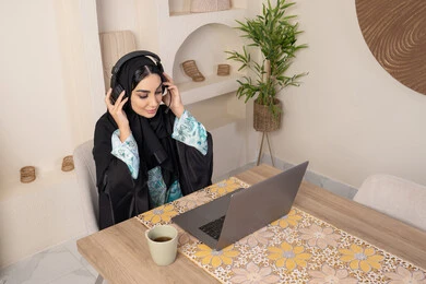 A calm morning atmosphere at home, enjoying drinking coffee, integrating modern technology with daily routine, a healthy lifestyle, a Saudi Arabian Gulf woman wearing an abaya and hijab sitting at a table at home using a laptop, an Emirati woman wearing headphones