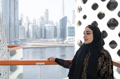 مشهد بانورامي لسيدة تستمتع بمشاهدة الأفق العمراني من شرفة مقهى حديث،شابة عربية خليجية اماراتية محجبة ترتدي عباءة سوداء مطرزة باللون الذهبي, الاستمتاع بالهواء الطلق