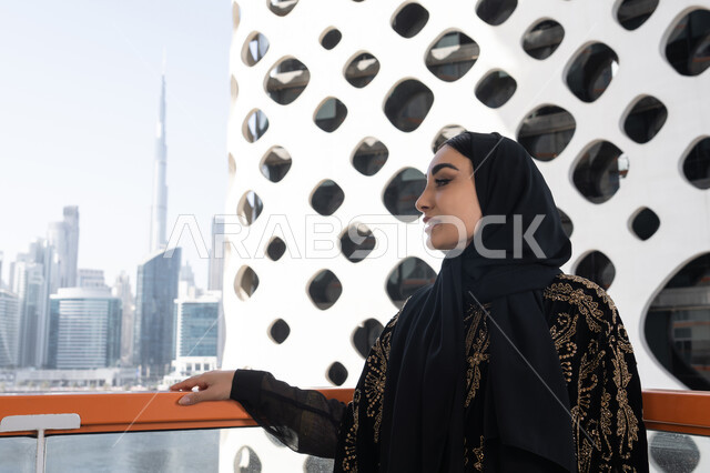 الهدوء والاستمتاع بالنظر من شرفة راقية, قضاء وقت ممتع في المقهى, شابة عربية خليجية اماراتية محجبة ترتدي عباءة سوداء مطرزة باللون الذهبي