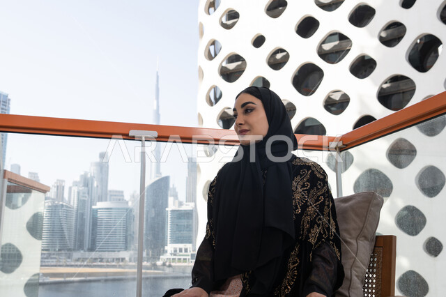 جلسة وناسة واستمتاع, لحظة صفاء راقية, شابة عربية خليجية اماراتية محجبة ترتدي عباءة سوداء مطرزة باللون الذهبي و تنظر من الشرفة, تصميم مقهى عصري