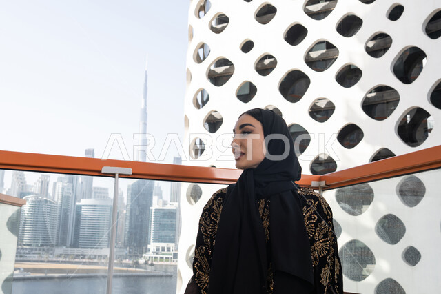 تصميم مقهى عصري و راقي، الهدوء والاستمتاع بالنظر من شرفة عالية,  شابة عربية خليجية اماراتية محجبة ترتدي عباءة سوداء مطرزة باللون الذهبي و تنظر بعيدا