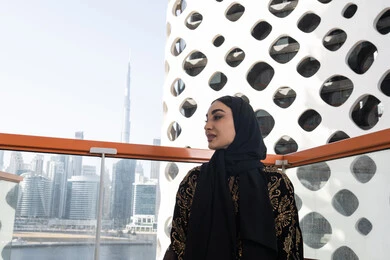 تصميم مقهى عصري و راقي، الهدوء والاستمتاع بالنظر من شرفة عالية,  شابة عربية خليجية اماراتية محجبة ترتدي عباءة سوداء مطرزة باللون الذهبي و تنظر بعيدا