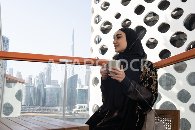 قهوة وإطلالة راقية,  قضاء وقت ممتع في المقهى, شابة عربية خليجية اماراتية محجبة ترتدي عباءة سوداء مطرزة باللون الذهبي و تنظر من الشرفة