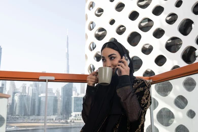 الإمساك بالهاتف المحمول والتواصل مع الأهل والأصدقاء, شابة عربية خليجية اماراتية محجبة مبتسمة ترتدي عباءة سوداء مطرزة باللون الذهبي, الاستمتاع بتناول مشروب ساخن