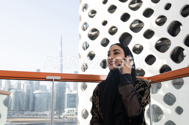 Communicating with the world easily, friendly conversations and dialogues, making a phone call using a mobile phone, a smiling veiled young Arab Gulf Emirati woman wearing a black abaya embroidered with gold