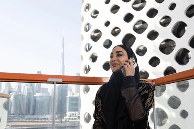 التواصل مع العالم بسهولة، محادثات وحوارات ودية، إجراء مكالمة هاتفية باستخدام الهاتف المحمول، شابة عربية خليجية اماراتية محجبة مبتسمة ترتدي عباءة سوداء مطرزة باللون الذهبي