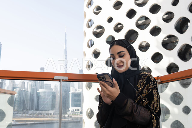 متابعة آخر المستجدات حول العالم, إيماءات الفرح والسرور، شابة عربية خليجية اماراتية محجبة مبتسمة ترتدي عباءة سوداء مطرزة باللون الذهبي و تجلس في مقهى راقي