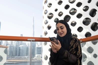 متابعة آخر المستجدات حول العالم, إيماءات الفرح والسرور، شابة عربية خليجية اماراتية محجبة مبتسمة ترتدي عباءة سوداء مطرزة باللون الذهبي و تجلس في مقهى راقي