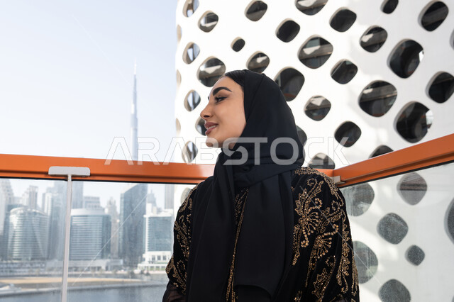 لحظة تأمل في مقهى مرتفع مطل على المدينة, الهدوء والاستمتاع بالنظر من شرفة راقية, شابة عربية خليجية اماراتية محجبة ترتدي عباءة سوداء مطرزة باللون الذهبي