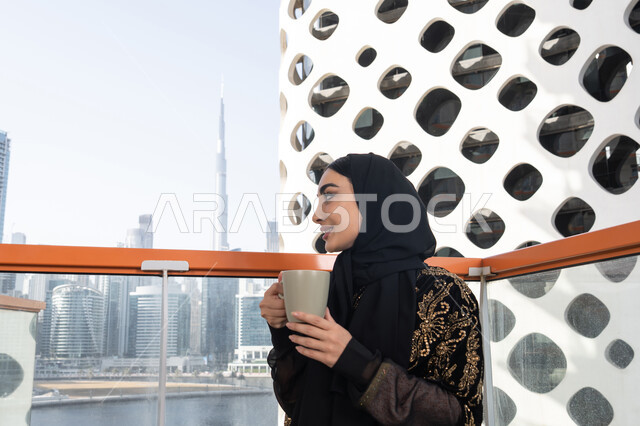 ابتسامة تعكس الرضا والارتياح, الاستمتاع بكوب من المشروب الدافئ, شابة عربية خليجية اماراتية محجبة ترتدي عباءة سوداء مطرزة باللون الذهبي