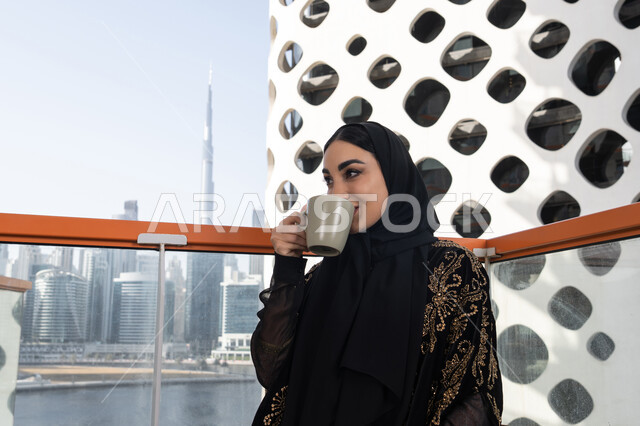 جلسة استرخاء مع فنجان قهوة, قضاء وقت ممتع في المقهى, شابة عربية خليجية اماراتية محجبة ترتدي عباءة سوداء مطرزة باللون الذهبي