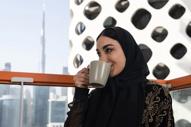 ارتشاف مشروب ساخن بابتسامة, شابة عربية خليجية اماراتية محجبة ترتدي عباءة سوداء مطرزة باللون الذهبي, إيماءات سعادة و سرور, مقهى مرتفع مطل على المدينة