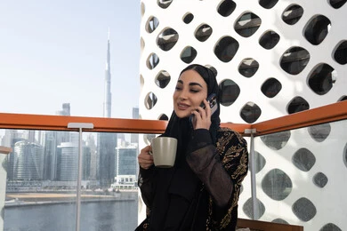 Hearing good news about the family's condition, making a phone call using a mobile phone, a smiling veiled young Arab Gulf Emirati woman wearing a black abaya embroidered with gold and holding a cup of hot drink