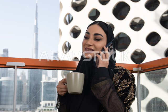 محادثات وحوارات ودية، إجراء مكالمة هاتفية باستخدام الهاتف المحمول، شابة عربية خليجية اماراتية محجبة مبتسمة ترتدي عباءة سوداء مطرزة باللون الذهبي و تحمل كوب من المشروب الدافئ