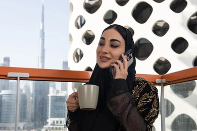 محادثات وحوارات ودية، إجراء مكالمة هاتفية باستخدام الهاتف المحمول، شابة عربية خليجية اماراتية محجبة مبتسمة ترتدي عباءة سوداء مطرزة باللون الذهبي و تحمل كوب من المشروب الدافئ