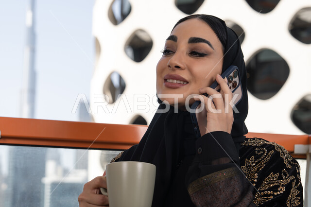 إيماءات الاندماج والسرور بالتواصل مع الأقارب عن بعد، مفهوم الجلوس والراحة، إجراء مكالمة هاتفية باستخدام الهاتف المحمول، شابة عربية خليجية اماراتية محجبة مبتسمة ترتدي عباءة سوداء مطرزة باللون الذهبي و تحمل كوب من المشروب الدافئ