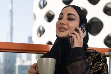 Gestures of integration and pleasure in communicating with relatives from a distance, the concept of sitting and resting, making a phone call using a mobile phone, a smiling veiled young Arab Gulf Emirati woman wearing a black abaya embroidered in gold