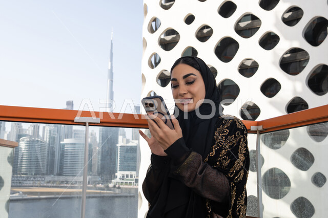 Reading a message on the screen, gestures of joy and happiness, a smiling veiled young Arab Gulf Emirati woman wearing a black abaya embroidered with gold, a modern café sitting and a view of skyscrapers