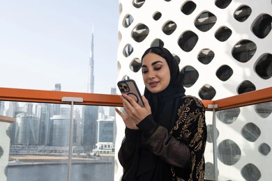قراءة رسالة على الشاشة, إيماءات الفرح والسرور, شابة عربية خليجية اماراتية محجبة مبتسمة ترتدي عباءة سوداء مطرزة باللون الذهبي, جلسة عصرية في مقهى حديث وإطلالة ناطحات السحاب