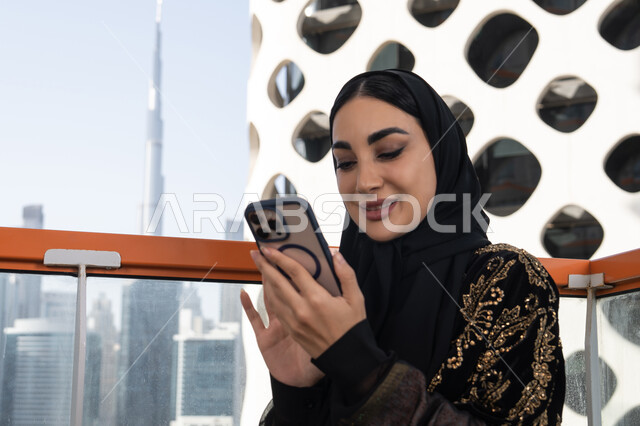تدوين الملاحظات على الهاتف المحمول, شابة عربية خليجية اماراتية محجبة مبتسمة ترتدي عباءة سوداء مطرزة باللون الذهبي, جلسة عصرية في مقهى حديث وإطلالة ناطحات السحاب