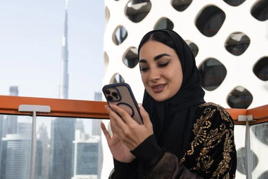 تدوين الملاحظات على الهاتف المحمول, شابة عربية خليجية اماراتية محجبة مبتسمة ترتدي عباءة سوداء مطرزة باللون الذهبي, جلسة عصرية في مقهى حديث وإطلالة ناطحات السحاب