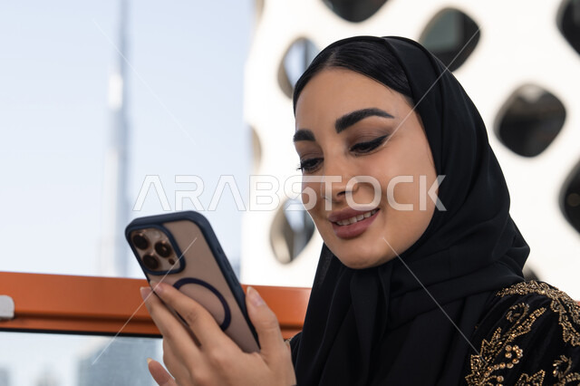 إيماءات الفرح والسرور لسماع الاخبار المفرحة، صورة مقربة لشابة عربية خليجية اماراتية محجبة مبتسمة ترتدي عباءة سوداء مطرزة باللون الذهبي, جلسة عصرية في مقهى حديث