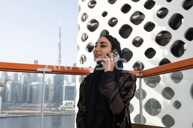 الرد والاجابة على الاتصالات الهاتفية عبر الهاتف المحمول، شابة عربية خليجية اماراتية محجبة مبتسمة ترتدي عباءة سوداء مطرزة باللون الذهبي, جلسة عصرية في مقهى حديث وإطلالة ناطحات السحاب
