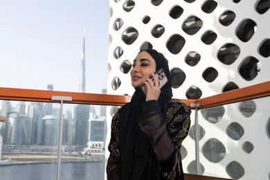 الرد والاجابة على الاتصالات الهاتفية عبر الهاتف المحمول، شابة عربية خليجية اماراتية محجبة مبتسمة ترتدي عباءة سوداء مطرزة باللون الذهبي, جلسة عصرية في مقهى حديث وإطلالة ناطحات السحاب