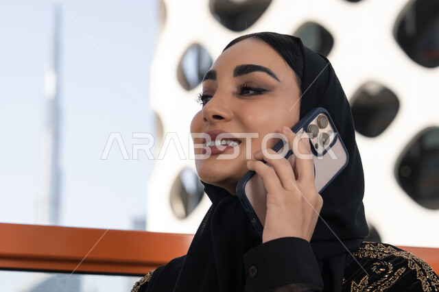 تعابير الاندماج والتركيز بالتحدث عبر الهاتف المحمول، قضاء اوقات ممتعة بتبادل الاخبار والأحاديث،  شابة عربية خليجية اماراتية محجبة مبتسمة ترتدي عباءة سوداء مطرزة باللون الذهبي