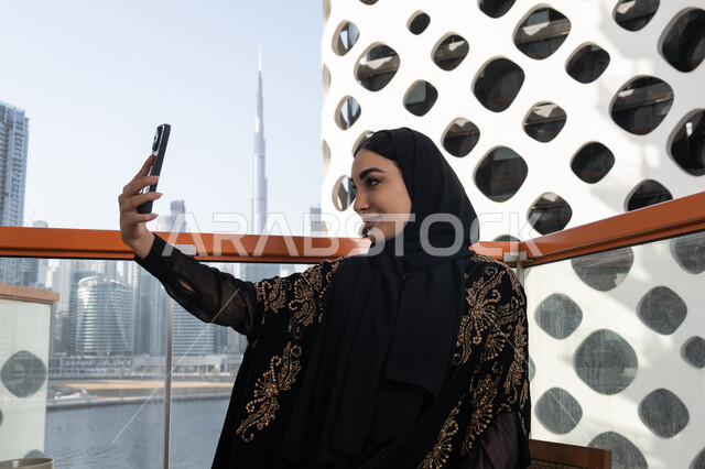 توثيق اللحظات السعيدة، إلتقاط صور سيلفي تذكارية بإستخدام الهاتف المحمول، إستخدام اجهزة تقنية حديثة، شابة عربية خليجية اماراتية محجبة مبتسمة ترتدي عباءة سوداء مطرزة باللون الذهبي