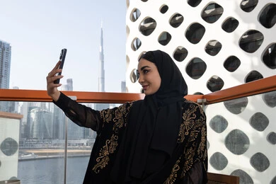 توثيق اللحظات السعيدة، إلتقاط صور سيلفي تذكارية بإستخدام الهاتف المحمول، إستخدام اجهزة تقنية حديثة، شابة عربية خليجية اماراتية محجبة مبتسمة ترتدي عباءة سوداء مطرزة باللون الذهبي
