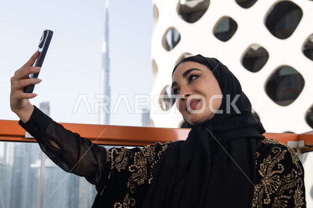 Taking souvenir selfies using a mobile phone, using modern technology devices, a smiling veiled young Arab Gulf Emirati woman wearing a black abaya embroidered in gold