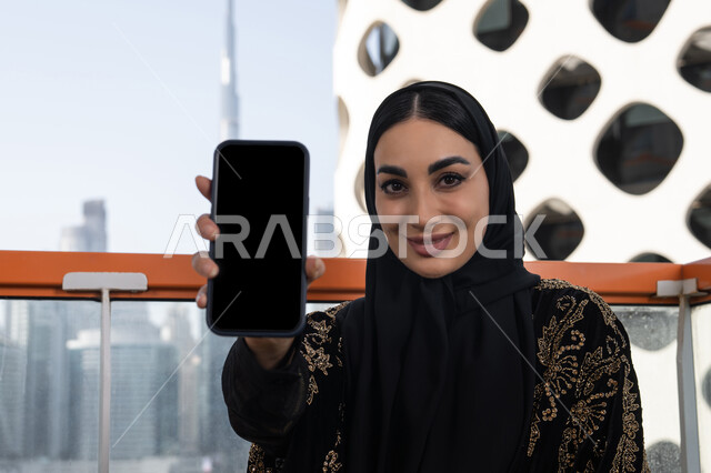 الاستجمام والتنزه في الهواء الطلق، عرض شاشة جوال سوداء فارغة، دمج التقنيات التكنولوجية الحديثة في الحياة اليومية، شابة عربية خليجية اماراتية محجبة مبتسمة ترتدي عباءة سوداء مطرزة باللون الذهبي