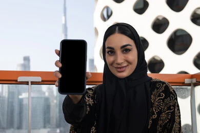 الاستجمام والتنزه في الهواء الطلق، عرض شاشة جوال سوداء فارغة، دمج التقنيات التكنولوجية الحديثة في الحياة اليومية، شابة عربية خليجية اماراتية محجبة مبتسمة ترتدي عباءة سوداء مطرزة باللون الذهبي