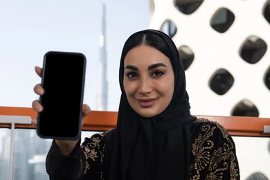 النظر إلى الكاميرا بإيماءات السعادة، عرض شاشة جوال سوداء فارغة، دمج التقنيات التكنولوجية الحديثة في الحياة اليومية، شابة عربية خليجية اماراتية محجبة مبتسمة ترتدي عباءة سوداء مطرزة باللون الذهبي