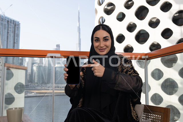 قضاء اوقات ممتعة بتبادل الاخبار والمحادثات عن بعد، عرض شاشة سوداء فارغة, شابة عربية خليجية اماراتية محجبة ترتدي عباءة سوداء مطرزة باللون الذهبي و تشير باصبع السبابة الي الهاتف