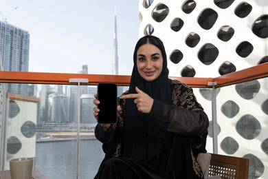 Having fun exchanging news and chatting remotely, a blank black screen is displayed, a young veiled Emirati Gulf Arab woman wearing a black abaya embroidered with gold and pointing her index finger at the phone