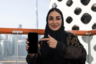 عرض شاشة جوال سوداء فارغة، دمج التقنيات التكنولوجية الحديثة في الحياة اليومية، شابة عربية خليجية اماراتية محجبة مبتسمة ترتدي عباءة سوداء مطرزة باللون الذهبي و تشير باصبع السبابة الي الهاتف