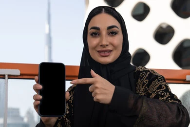حياة عصرية بلمسة تكنولوجية, شابة عربية خليجية اماراتية محجبة مبتسمة ترتدي عباءة سوداء مطرزة باللون الذهبي و تشير باصبع السبابة الي الهاتف, عرض شاشة سوداء فارغة
