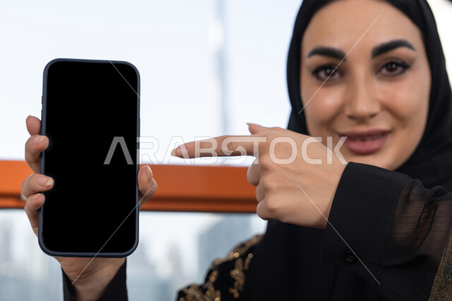 Close-up of a blank black screen display, using a modern advanced technical device, a young veiled Arab Gulf Emirati woman wearing a black abaya embroidered in gold and pointing her index finger at the phone