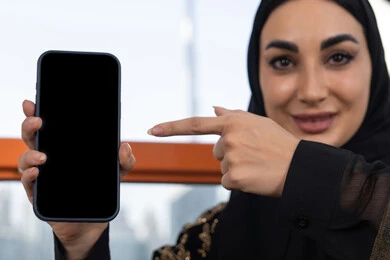 صورة مقربة لعرض شاشة سوداء فارغة, استخدام جهاز تقني حديث متطور، شابة عربية خليجية اماراتية محجبة ترتدي عباءة سوداء مطرزة باللون الذهبي و تشير باصبع السبابة الي الهاتف