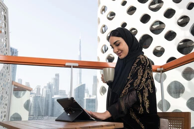 قهوة وإطلالة راقية,  شابة عربية خليجية اماراتية محجبة ترتدي عباءة سوداء مطرزة باللون الذهبي و تقف في مقهى مرتفع مطل على المدينة, الاستمتاع بتناول المشروب الساخن