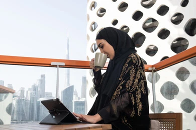 مساحة يلتقي فيها الإلهام بالتقنية, شابة عربية خليجية اماراتية محجبة ترتدي عباءة سوداء مطرزة باللون الذهبي و تقف في مقهى مرتفع مطل على المدينة, الاستمتاع بتناول المشروب الساخن