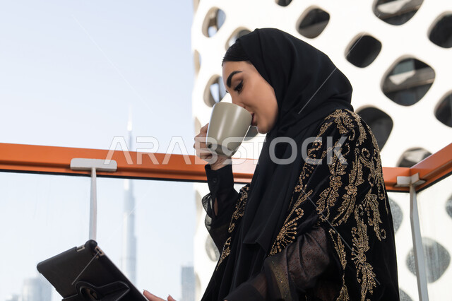 لحظة استراحة مع فنجان دافئ مع متابعة الأعمال عبر الجهاز اللوحي, شابة عربية خليجية اماراتية محجبة ترتدي عباءة سوداء مطرزة باللون الذهبي و تقف في مقهى مرتفع مطل على المدينة, الاستمتاع بتناول المشروب الساخن
