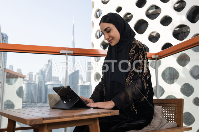 مشهد جانبي لسيدة  تقرأ من جهازها اللوحي أثناء وقوفها في مقهى حديث الطراز, شابة عربية خليجية اماراتية محجبة ترتدي عباءة سوداء مطرزة باللون الذهبي